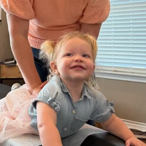 Child on a treatment table while another person stands behind, with a window and blinds in the background.